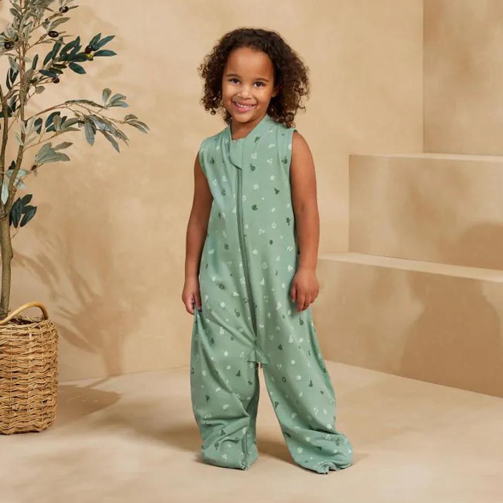 Child wearing a green patterned outfit standing in front of a beige wall with a plant.
