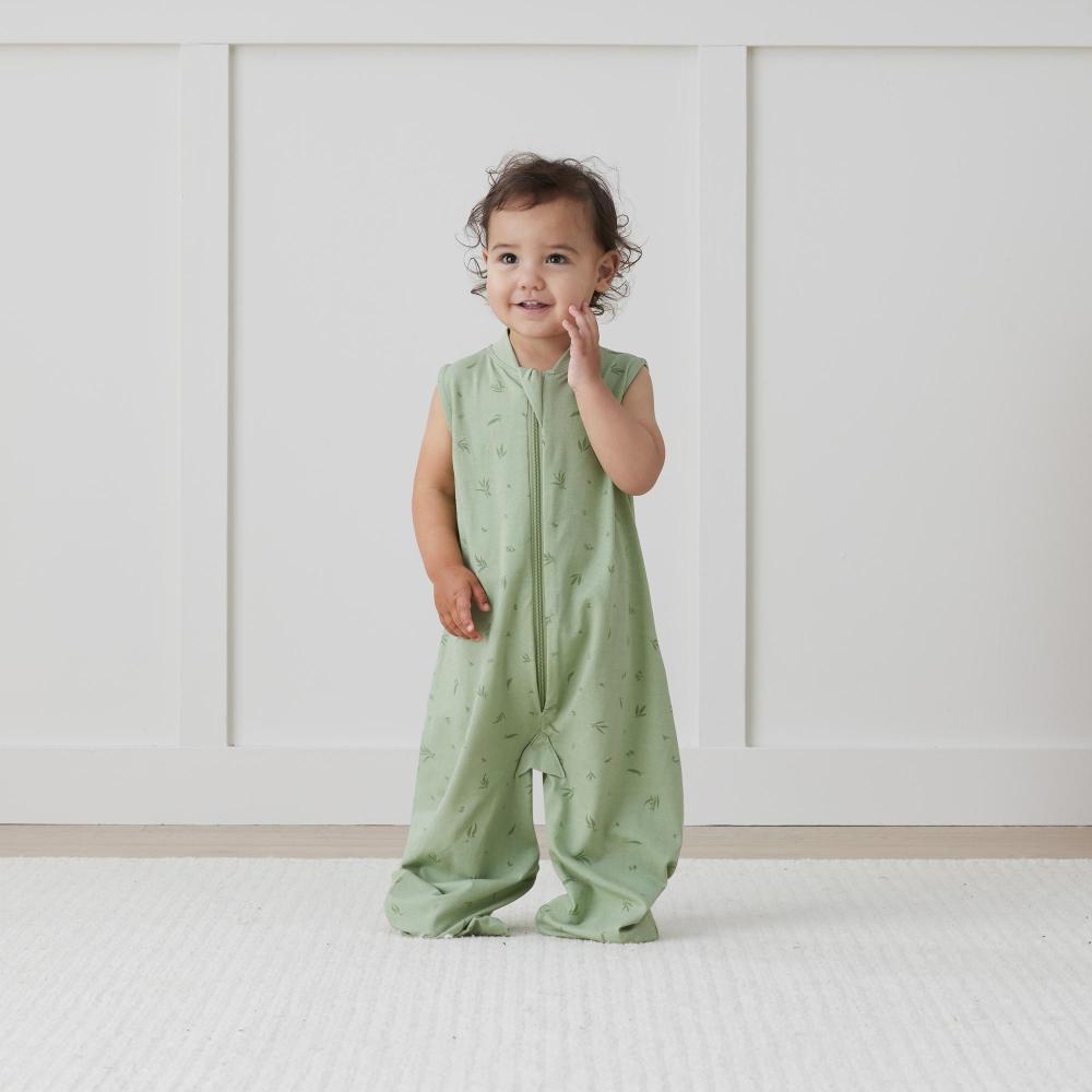 Child wearing a green sleeveless jumpsuit standing on a white floor with a light-colored wall in the background.