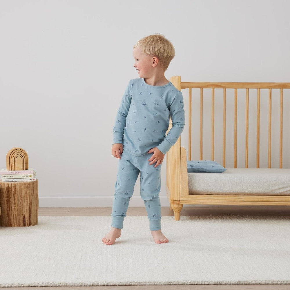 Child wearing blue pajamas standing in a bedroom with a crib and wooden toys.