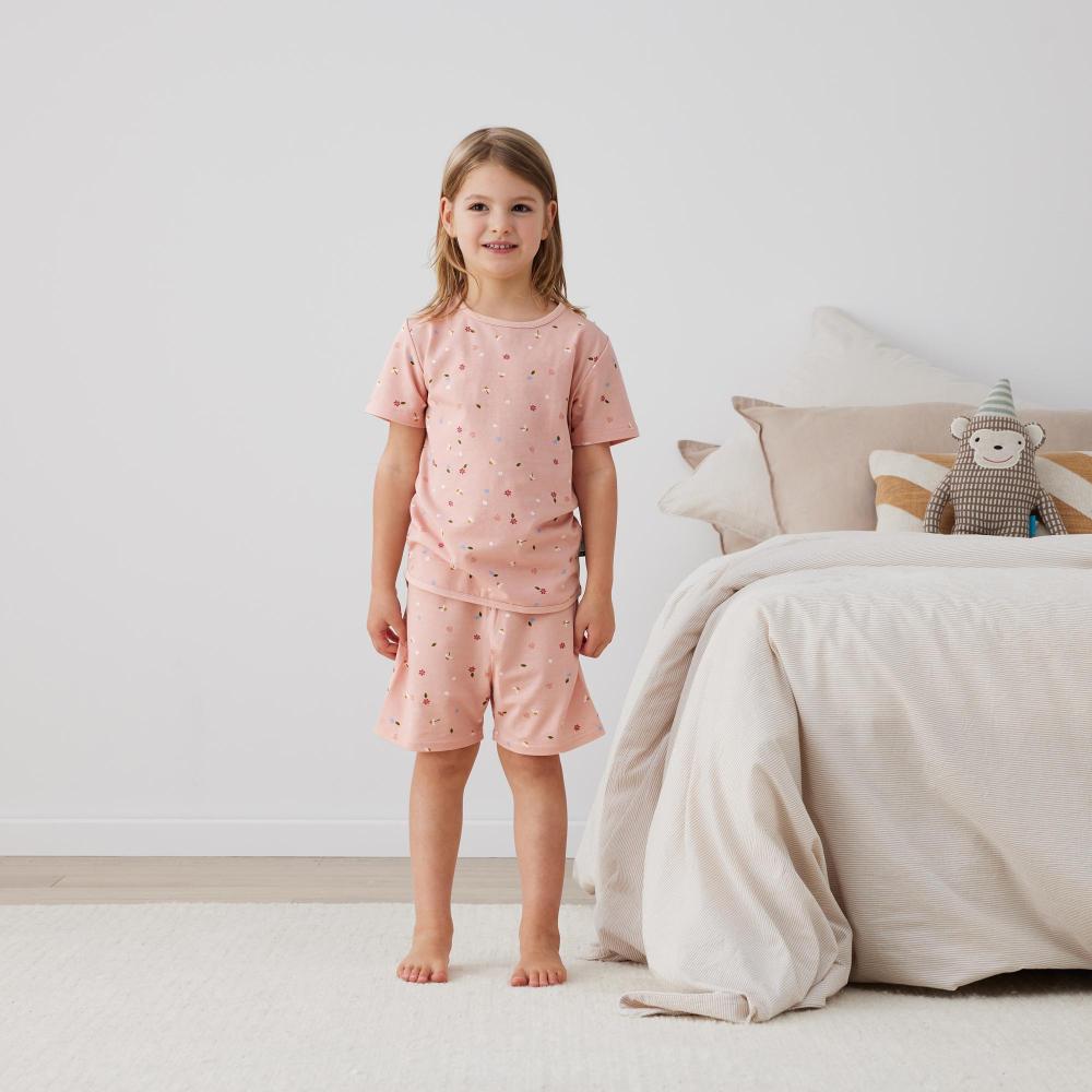 Young girl in pink pajamas standing in a bedroom with a bed and teddy bear in the background.