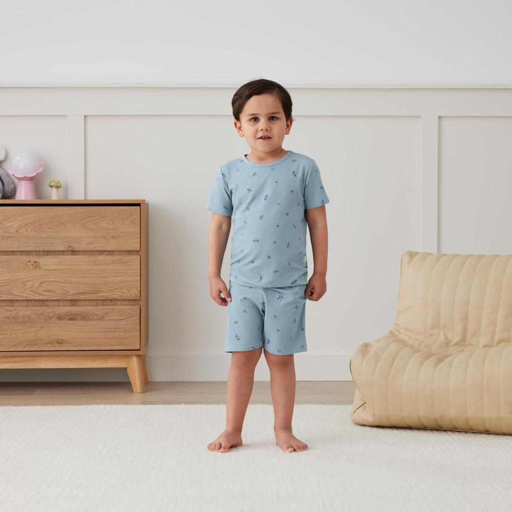 Child wearing light blue shortie pajamas standing in a room with wooden furniture and a beige bean bag chair.
