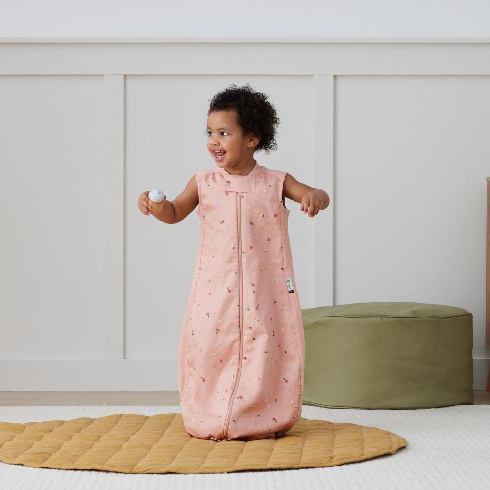 Child wearing a pink sleeveless sleep sack with a pattern, standing on a yellow mat in a room.