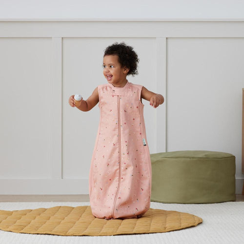 Child wearing a pink sleeping bag with a pattern, standing on a yellow mat in a room.