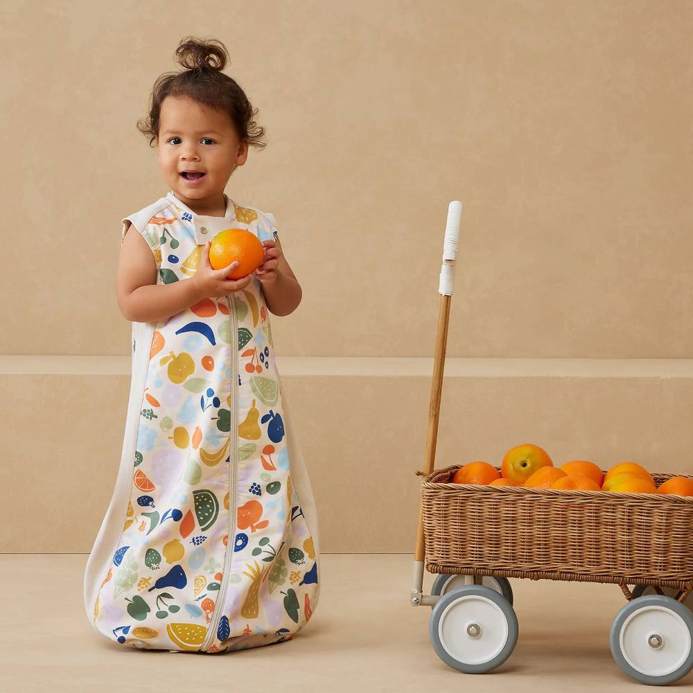 Child in a colorful sleep sack holding an orange next to a cart with more oranges on a beige background