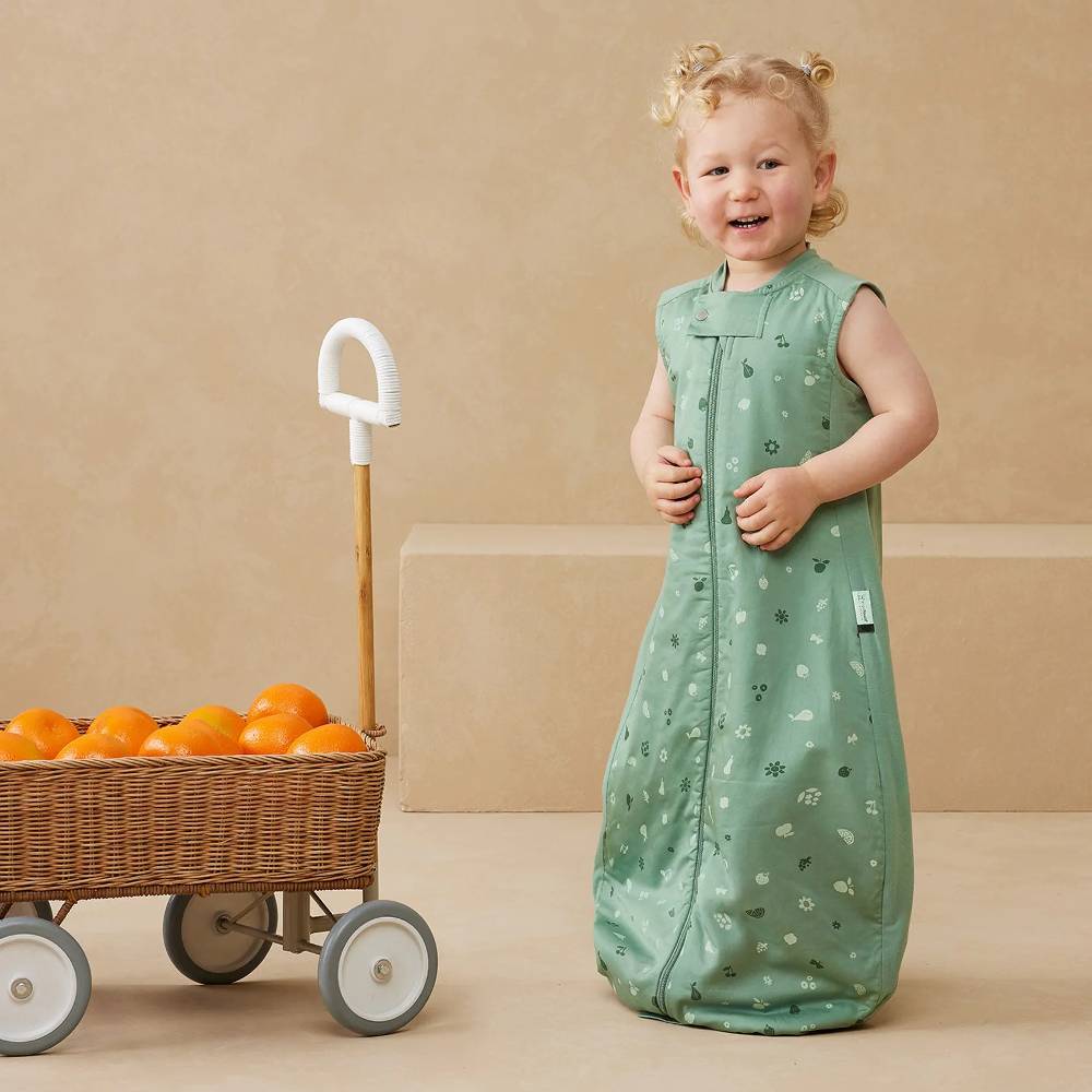 Child in a green sleeveless sleep suit standing next to a cart with oranges on a beige background