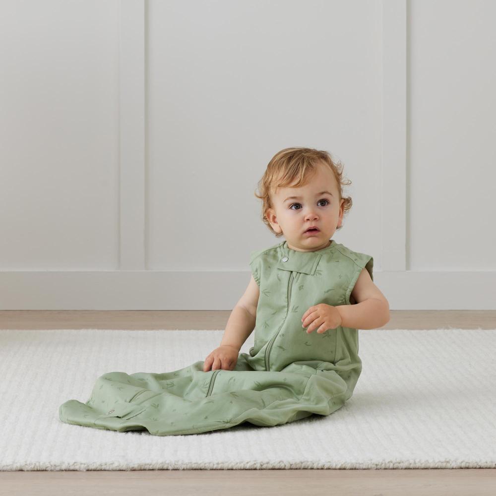 Baby in a green outfit sitting on a white rug with a white paneled wall background