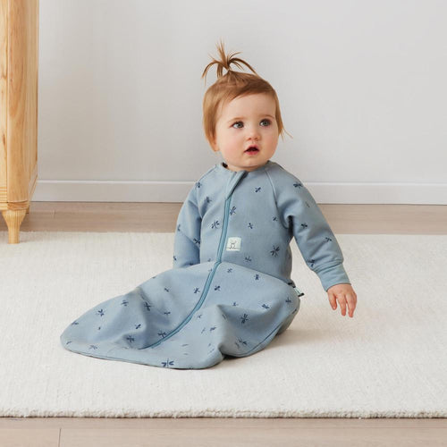 Baby in a blue sleep bag sitting on a white rug.