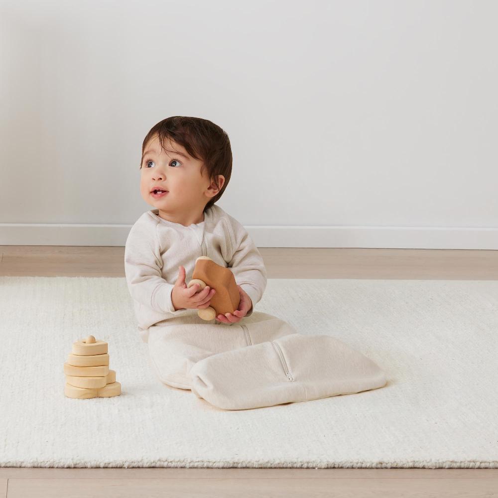 Baby sitting on a white rug in a sleeping bag holding a wooden toy, with another wooden toy stack nearby.