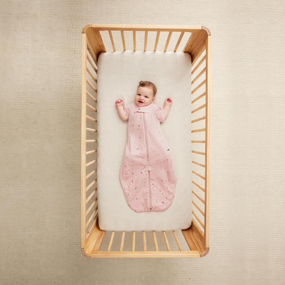 Baby in a pink outfit lying in a wooden crib with a beige wall background