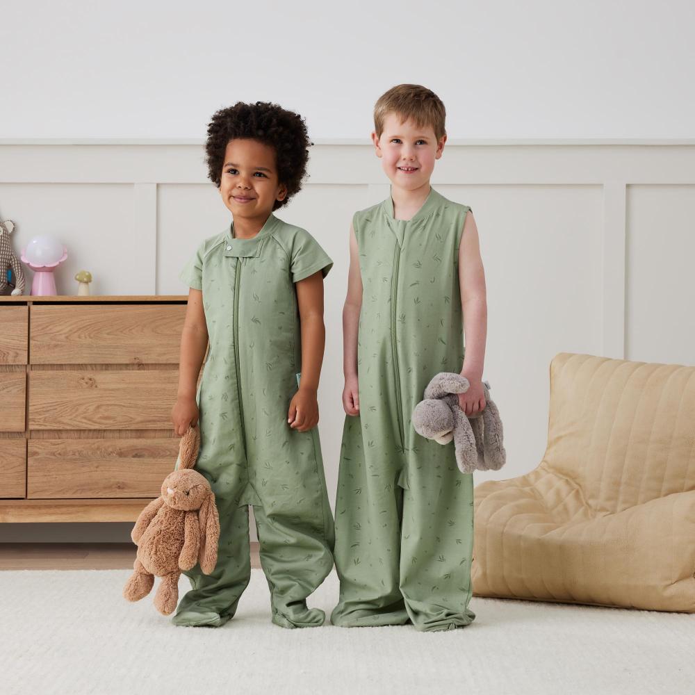 Two children wearing green sleeveless sleepsuits standing in a room with a wooden dresser and beige chair.