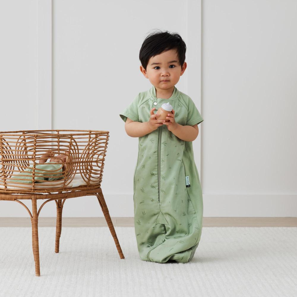 Child wearing a green baby sleepsuit in a minimalistic room with a wicker chair.
