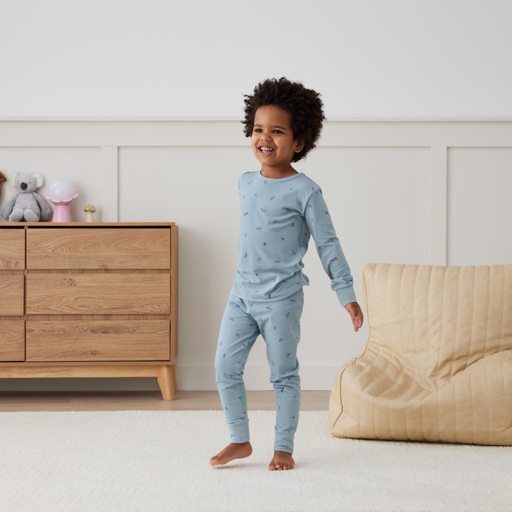 Child in light blue pajamas standing in a bedroom with wooden dresser and beige sofa.