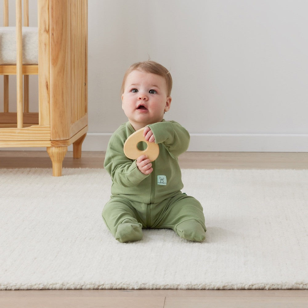 Baby in a green onesie holding a wooden toy in a nursery setting.