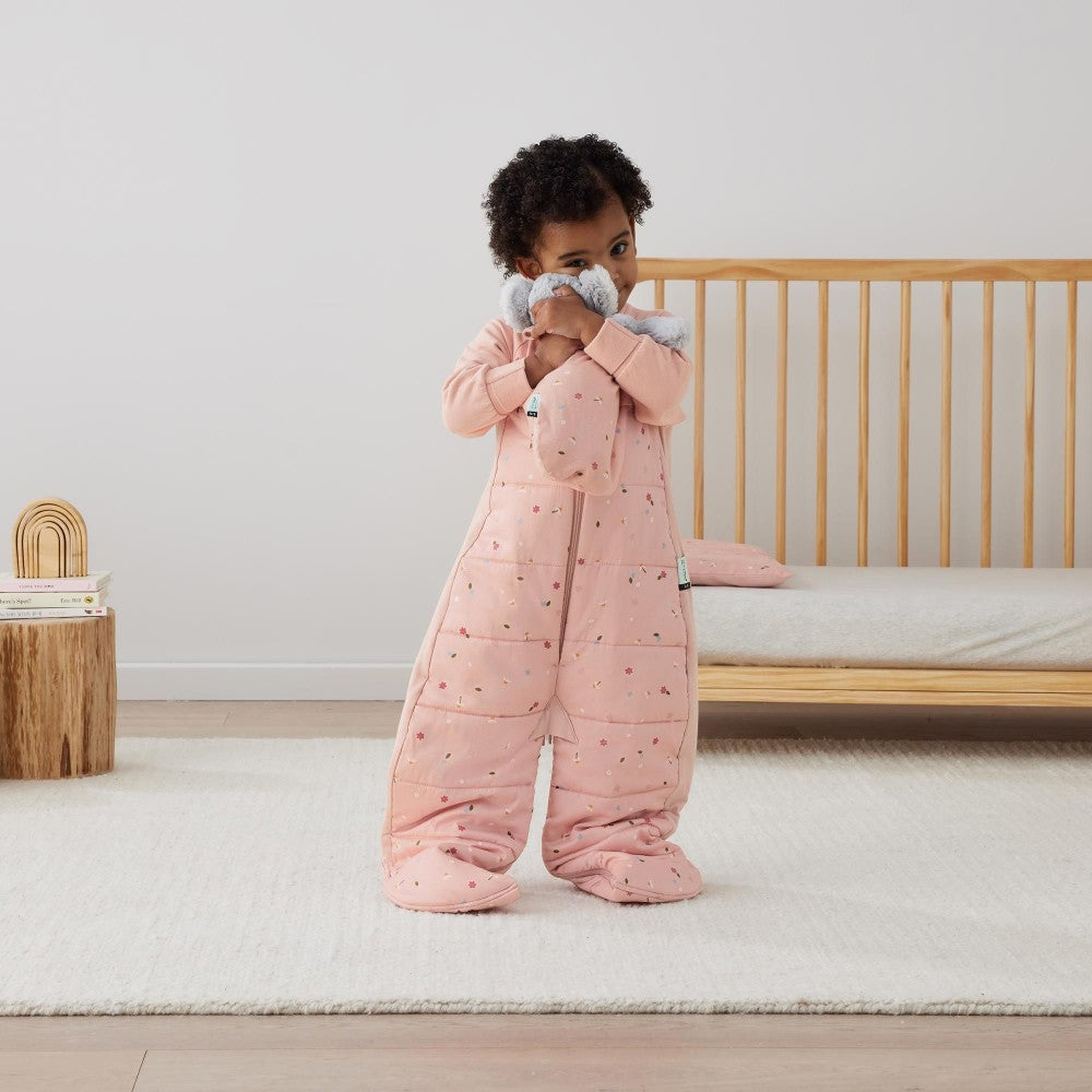 Child wearing a pink sleepsuit standing in a room with a crib and wooden toy.