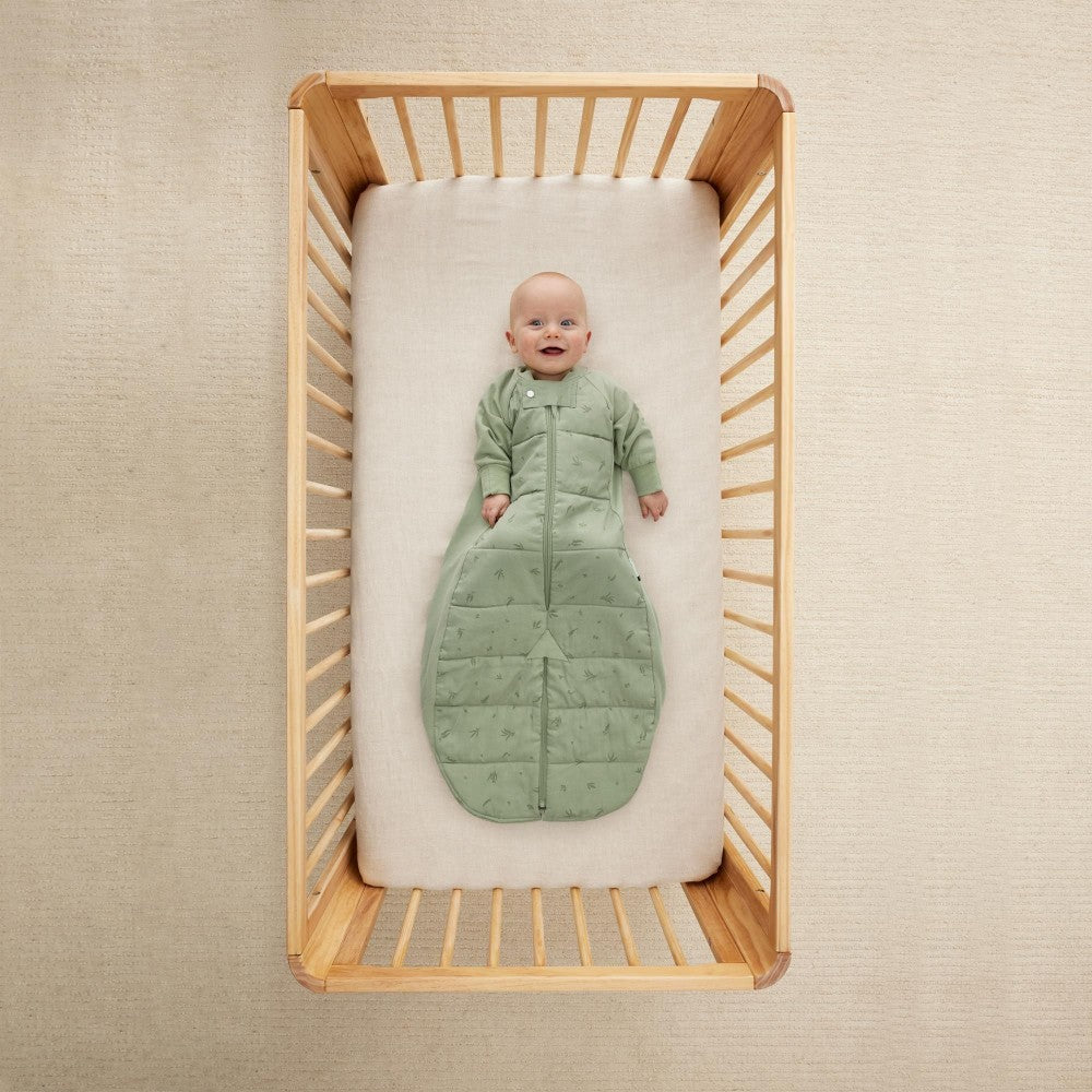Baby in a green sleeping bag lying in a wooden crib with a beige wall background