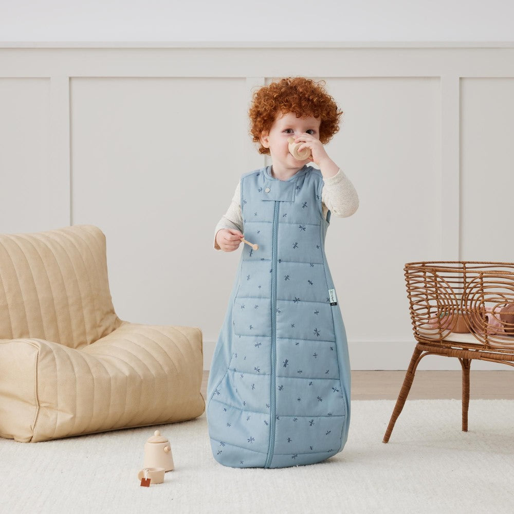 Child wearing a blue quilted sleep sack in a room with beige furniture and a white wall.
