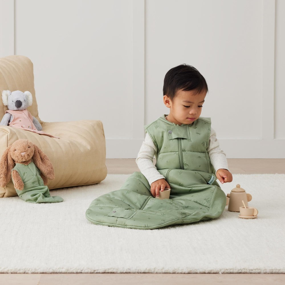 Child in a green outfit sitting on a white rug with toys around