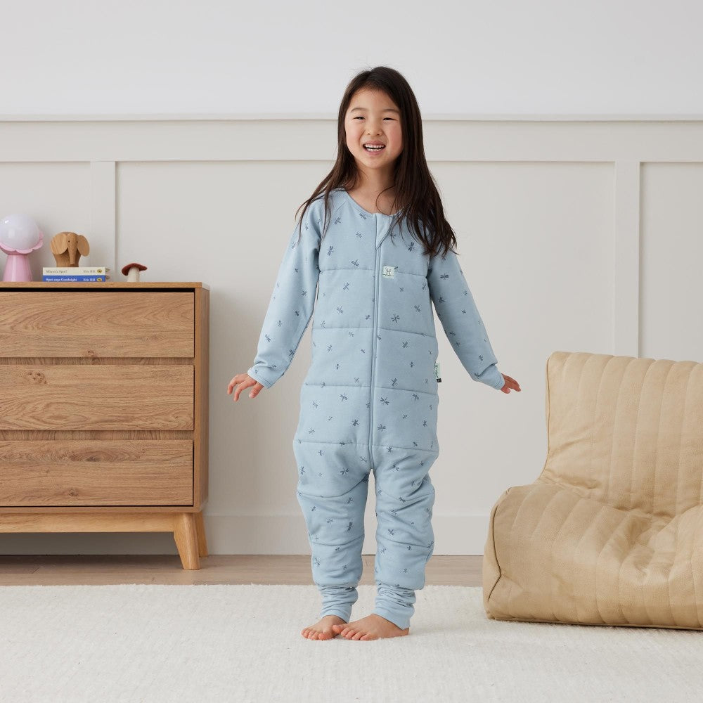 Child wearing a light blue onesie with a pattern in a room with a wooden dresser and beige chair.