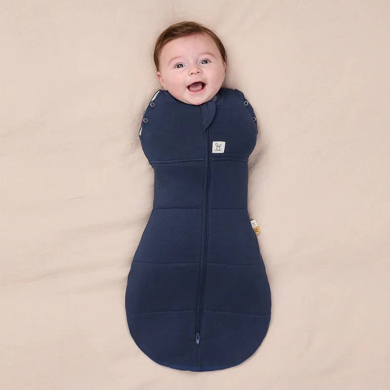 Baby lying on a beige surface wearing a navy blue swaddle sleeping bag