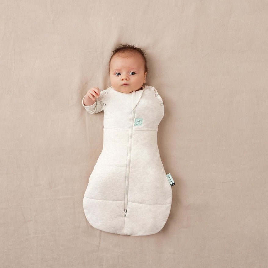 Baby lying on a beige surface wearing a cream sleeping bag with a small green tag.