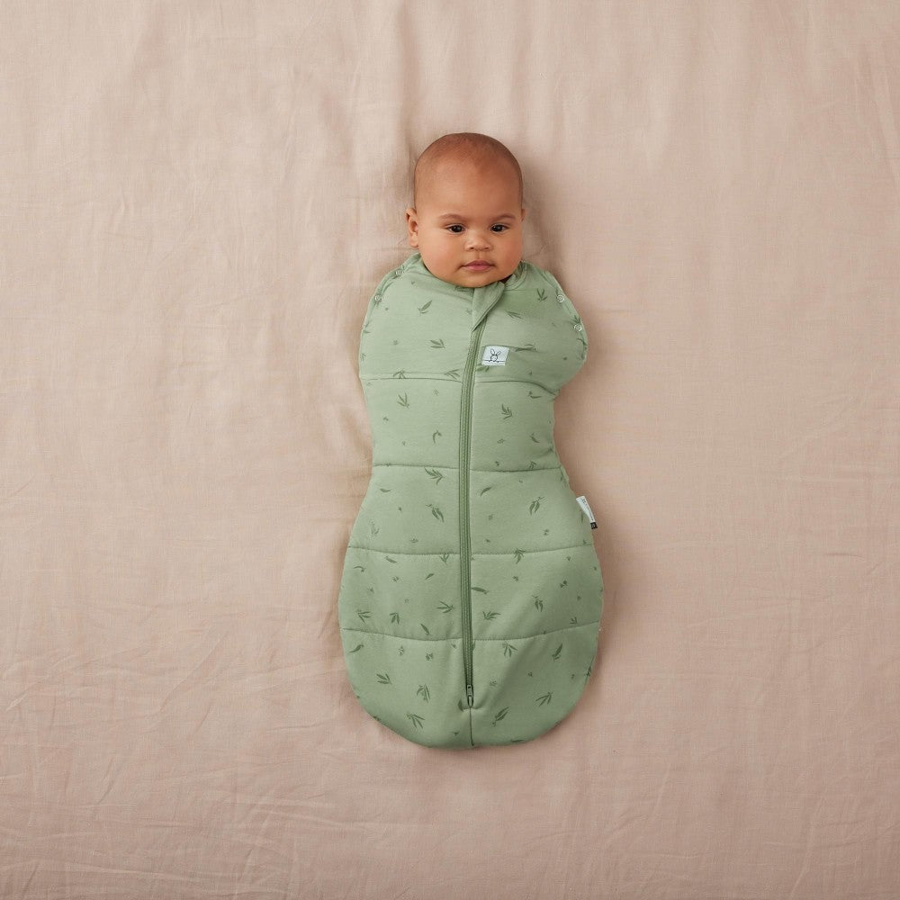 Baby wrapped in a green sleeping bag on a beige surface