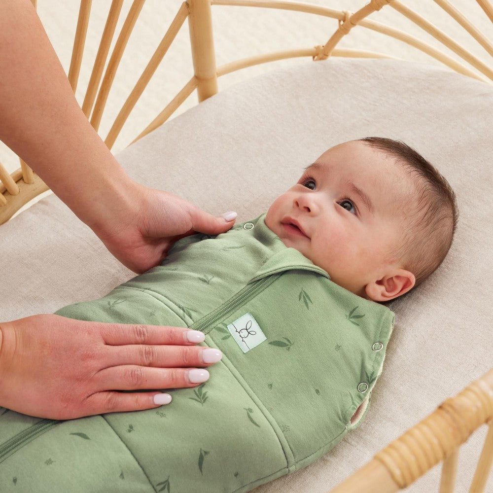 Baby in a green swaddling bag being adjusted by a hand in a crib