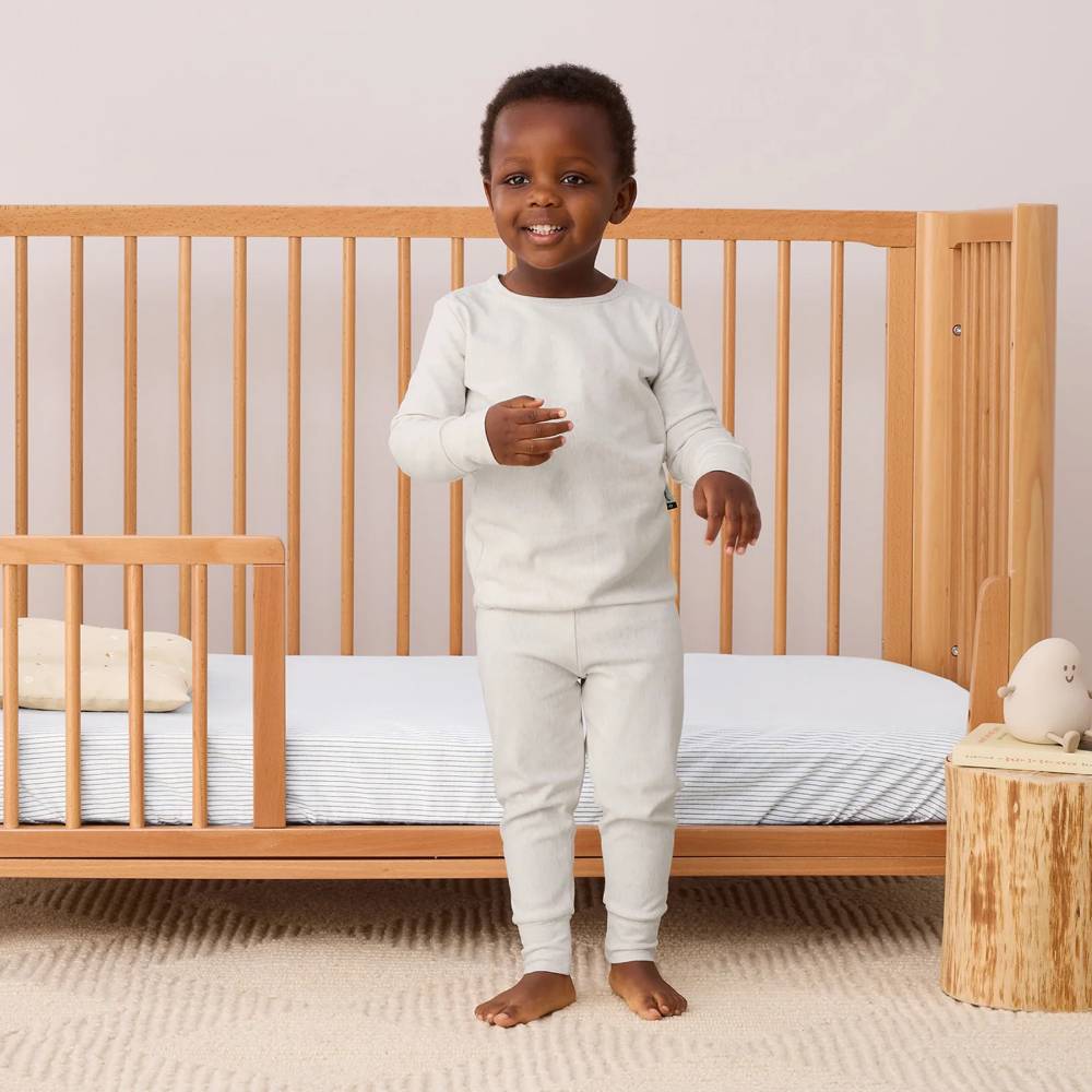Child in oatmeal pajamas standing in front of a wooden crib with a neutral background