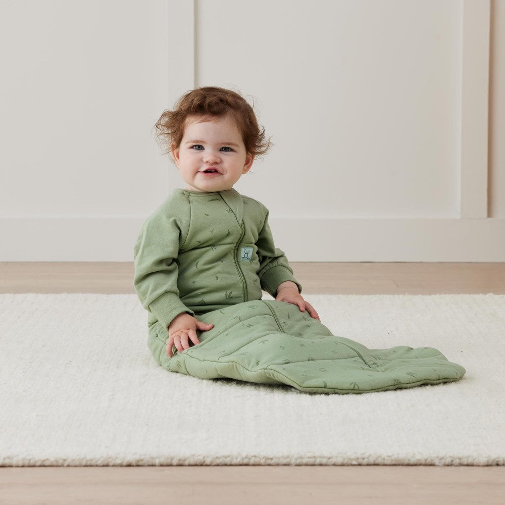 Baby in a green sleeping bag sitting on a white floor.