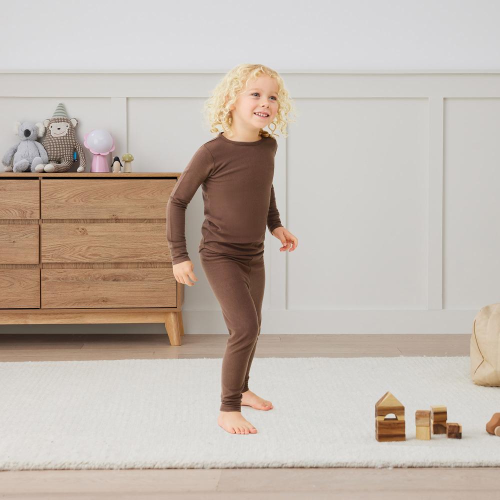 Child in brown pajamas standing in a room with wooden furniture and toys.