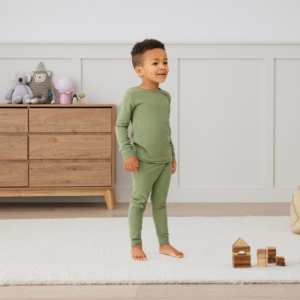Child wearing green pajamas standing in a bedroom with wooden dresser and toys.
