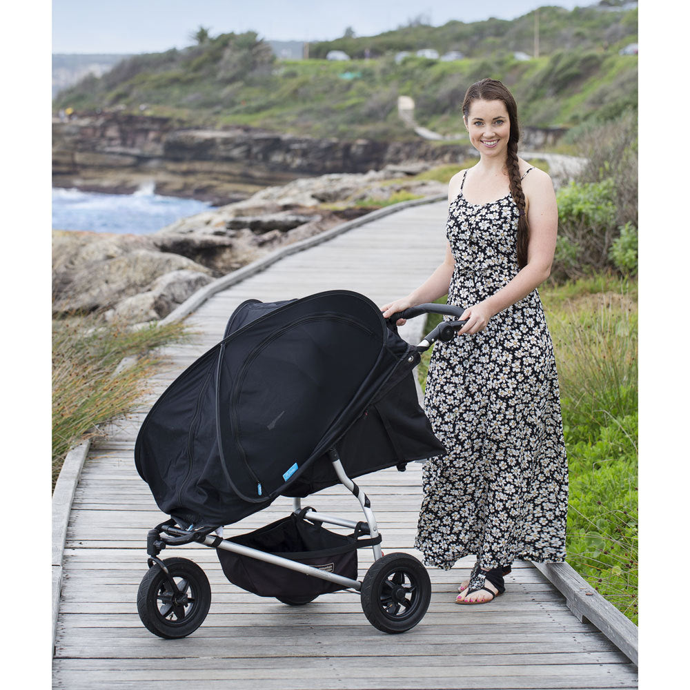 A woman in a black and white floral dress pushes a black stroller with a Cozi-Go Shade cover along a wooden boardwalk by the ocean, with rocky cliffs and green vegetation in the background.
