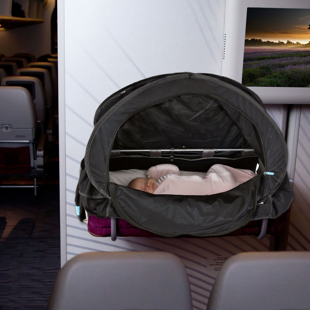 A baby sleeping in a dark gray pop-up bassinet set up on an airplane seat, with the airplane cabin using the Cozi Go Bassinet cover