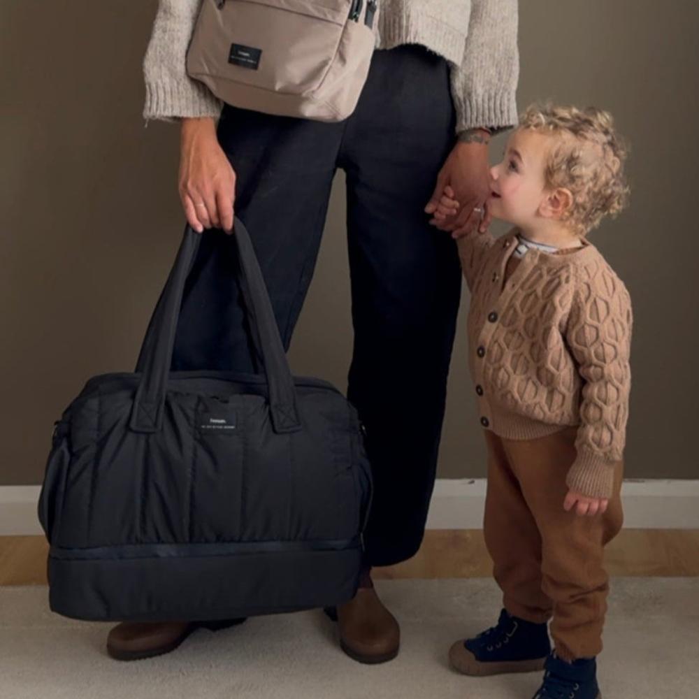 Person holding a navy duffel bag with a child in a beige sweater and brown pants standing next to them.