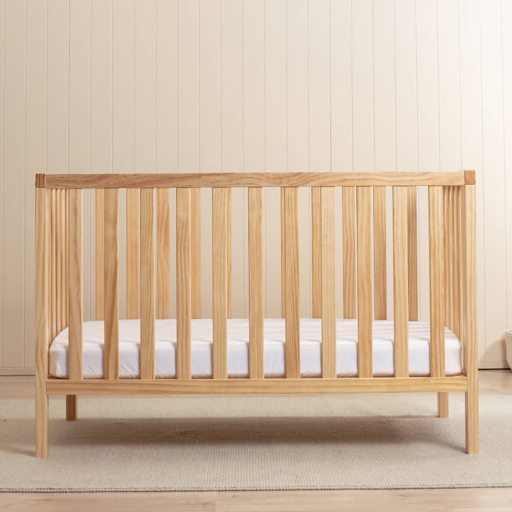 A light natural wood convertible crib or toddler bed with vertical slats, shown with a white mattress in a bright, neutral nursery setting.