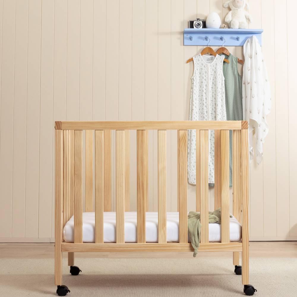 A light natural wood baby crib with vertical slats and black casters, shown in a styled nursery setting with a white mattress, a small green throw, and a blue wall-mounted coat rack with hanging baby clothes in the background.