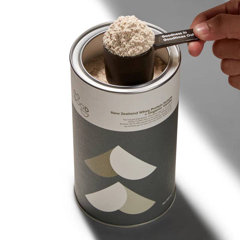 A hand holds a scoop of beige powder over an open cylindrical canister labeled 'New Zealand Whey Protein Powder' 