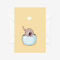 A yellow greeting card with a cartoon baby kiwi hatching out of an egg. There a white envelope behind it on a white background. 