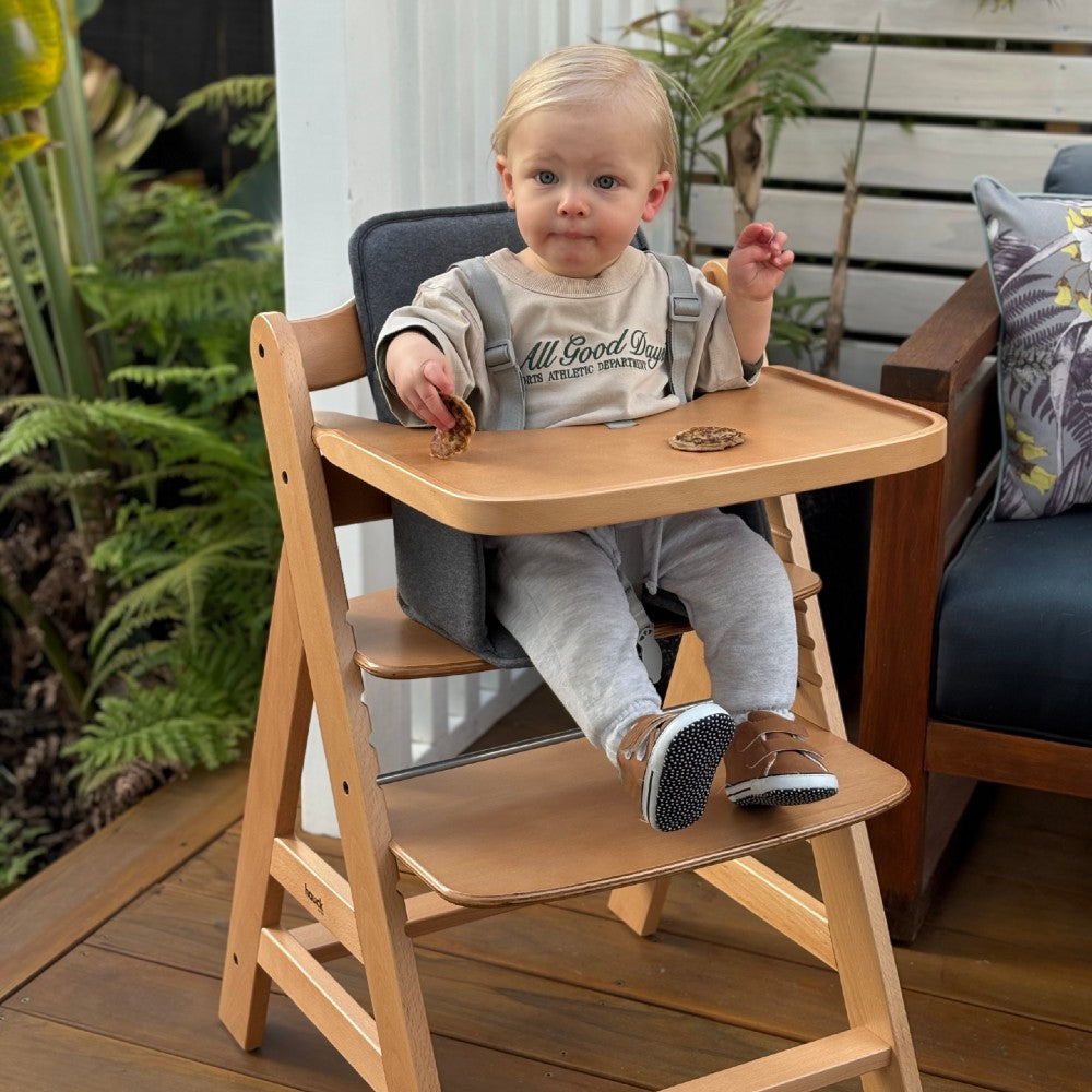 Child sitting in a Hauck wooden high chair outdoors with plants in the background