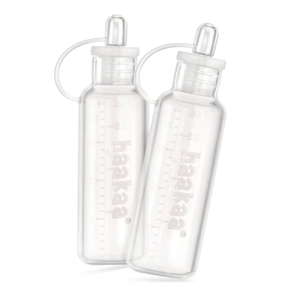 Two clear baby colostrum collectors with caps on a white background, featuring the brand 'Haakaa'.