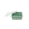 A silicone ice cube tray in sage green with six compartments and a translucent white lid resting at an angle on top, set against a plain white background.