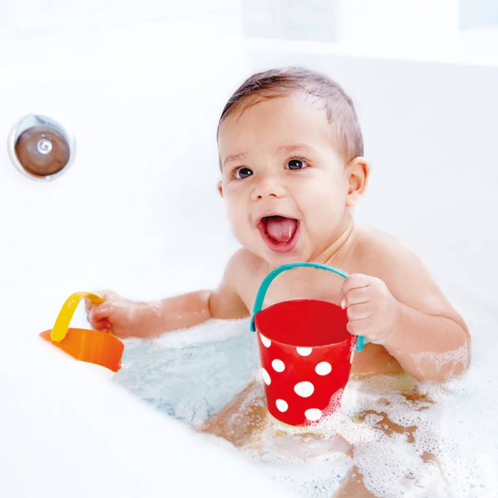 Baby playing with Hape bath buckets in bathtub against white background.