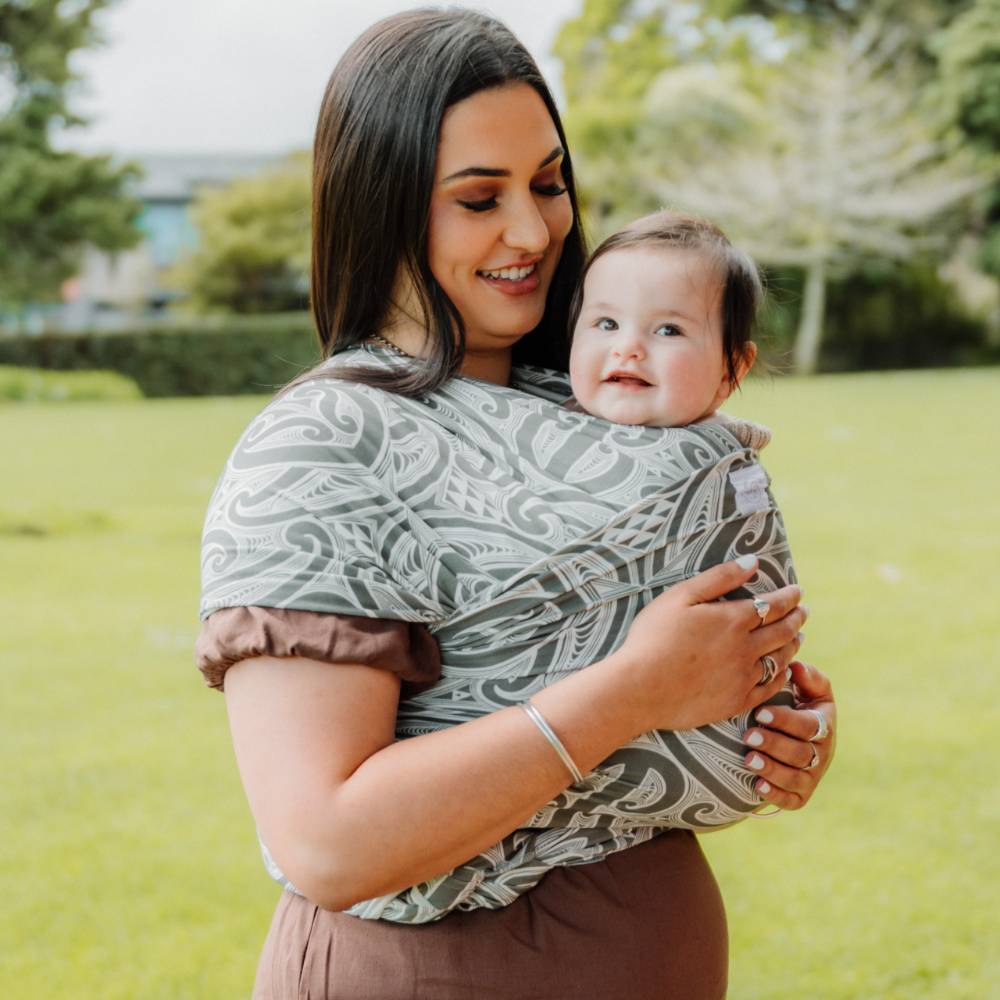 A woman outdoors holding a baby in a wrap-style baby carrier. The carrier has a gray and white Okooko Pepi pattern with 