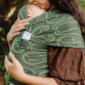 A close-up lifestyle shot of a woman wearing a Hiwa Maori print green and brown baby wrap carrier with a sleeping baby tucked inside, outdoors with a blurred natural background.