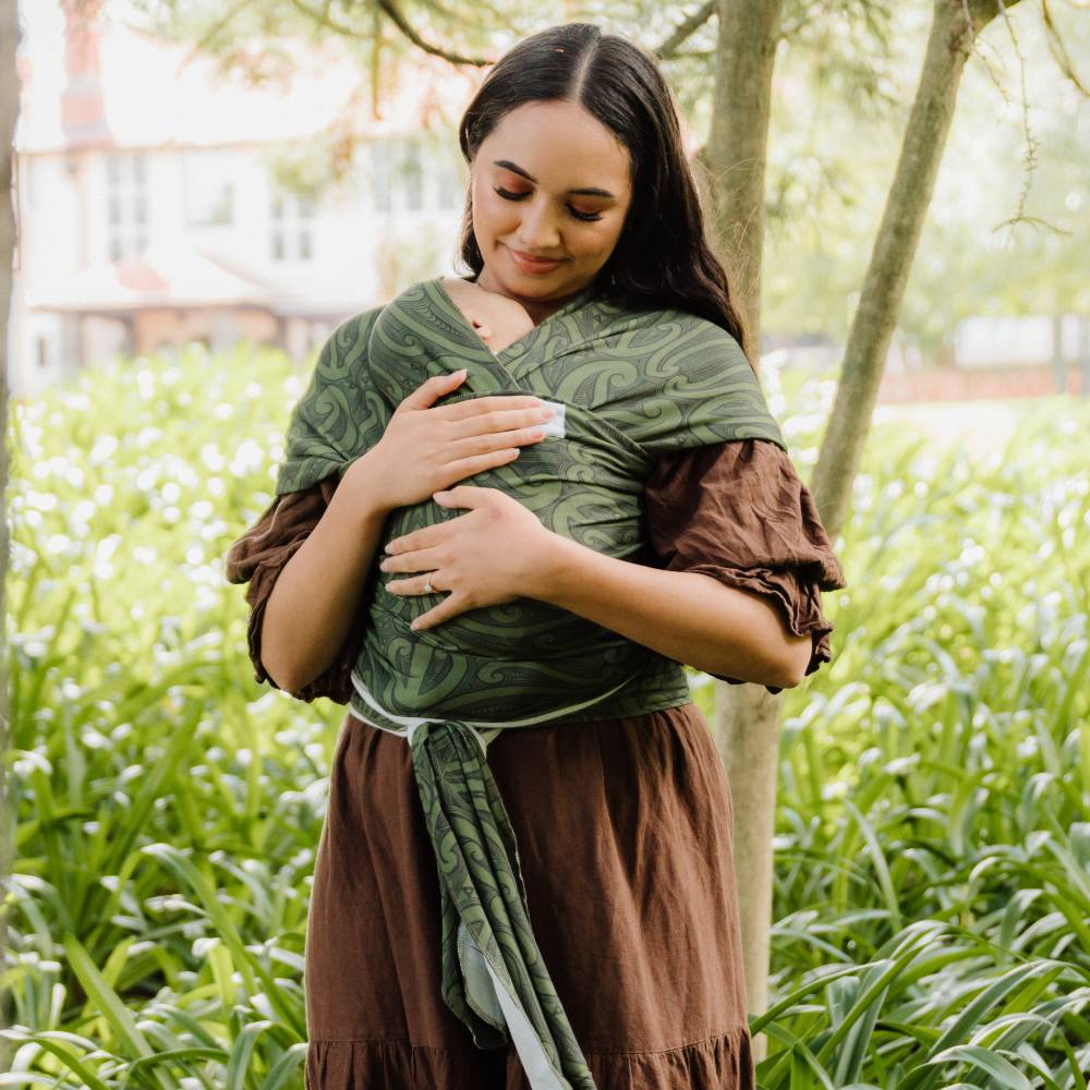 A woman outdoors wearing a brown dress and a green patterned baby wrap or shawl draped over her torso, standing in a grassy area with trees and a blurred building in the background.