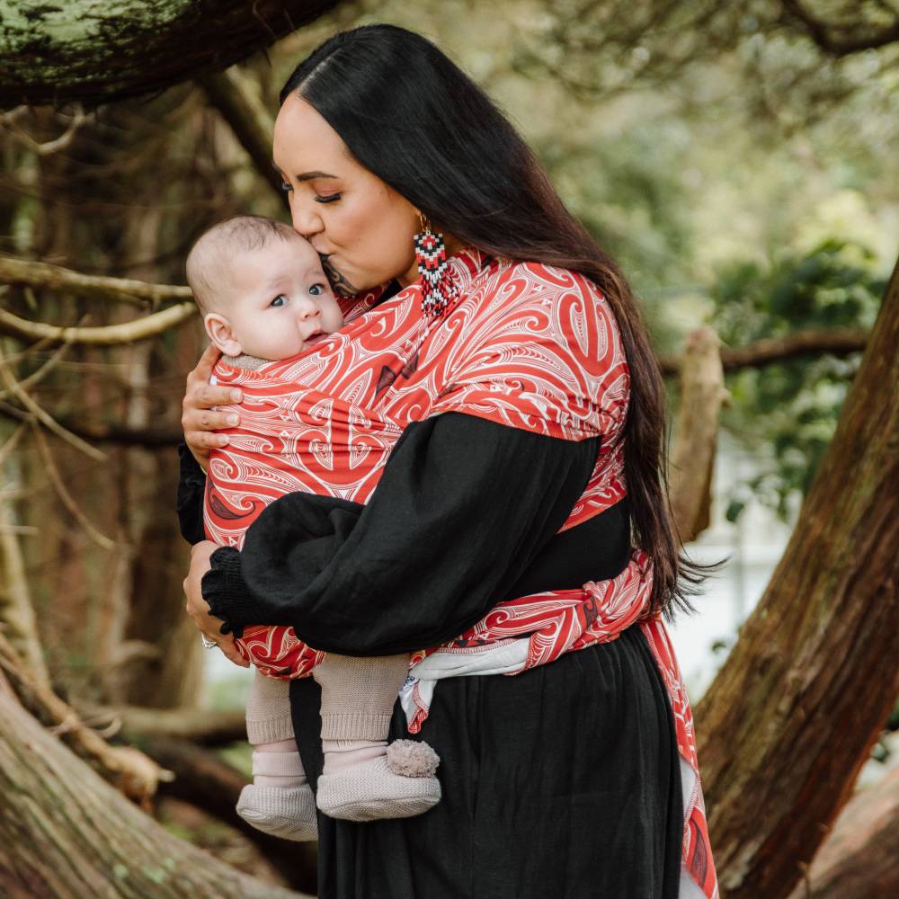 A woman outdoors carrying a baby in a red and white Maori patterned wrap-style baby carrier dress, with a black long-sleeve top underneath.