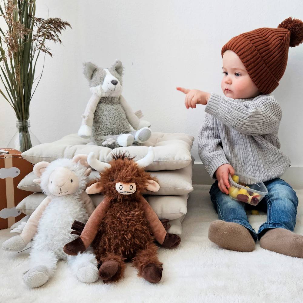 A baby sitting on the floor pointing to his soft toys - Happy Horse Cow Cody, Sheep and Husky Hunter. 
