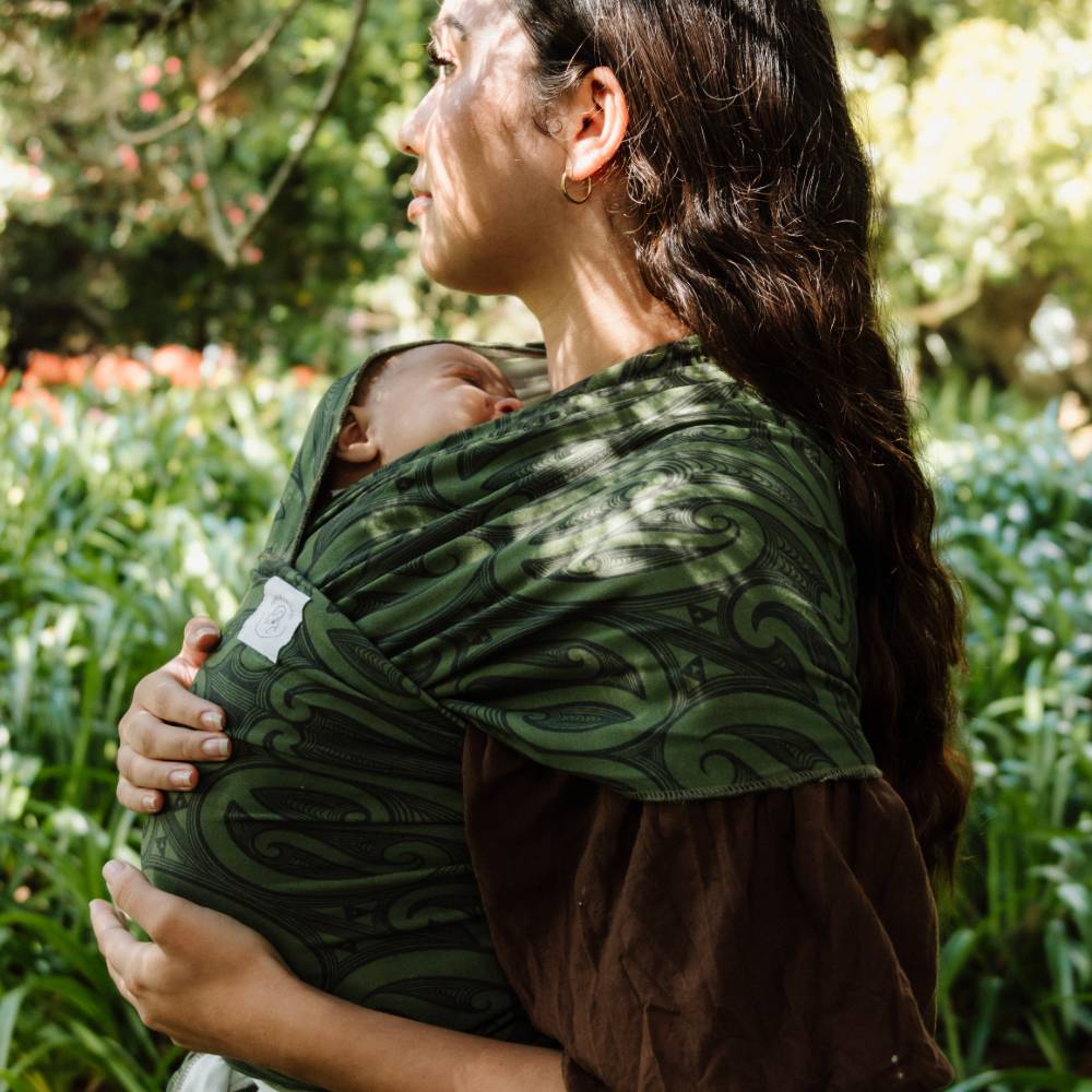 A woman outdoors wearing a dark green New Zealand patterned baby wrap carrier with a sleeping newborn baby tucked inside, set against a blurred green garden background.