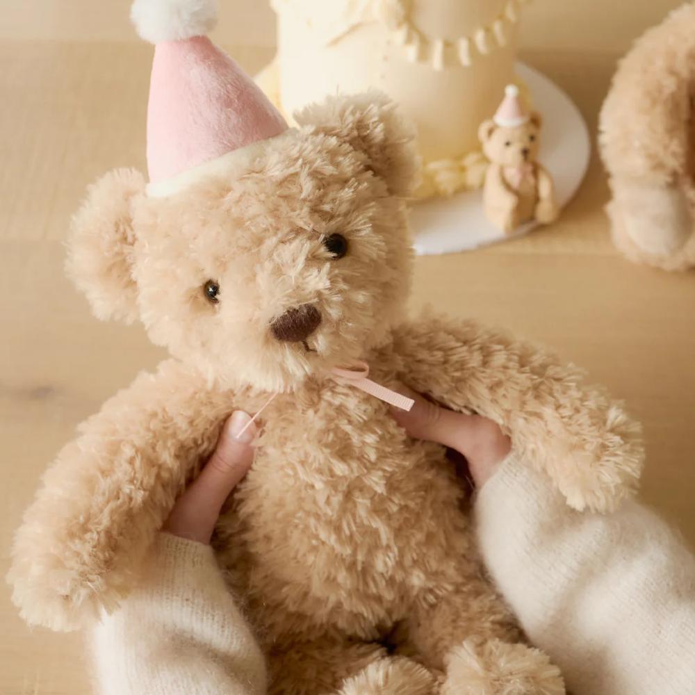 Brown Jamie Kay bear soft toy with pink hat being held.
