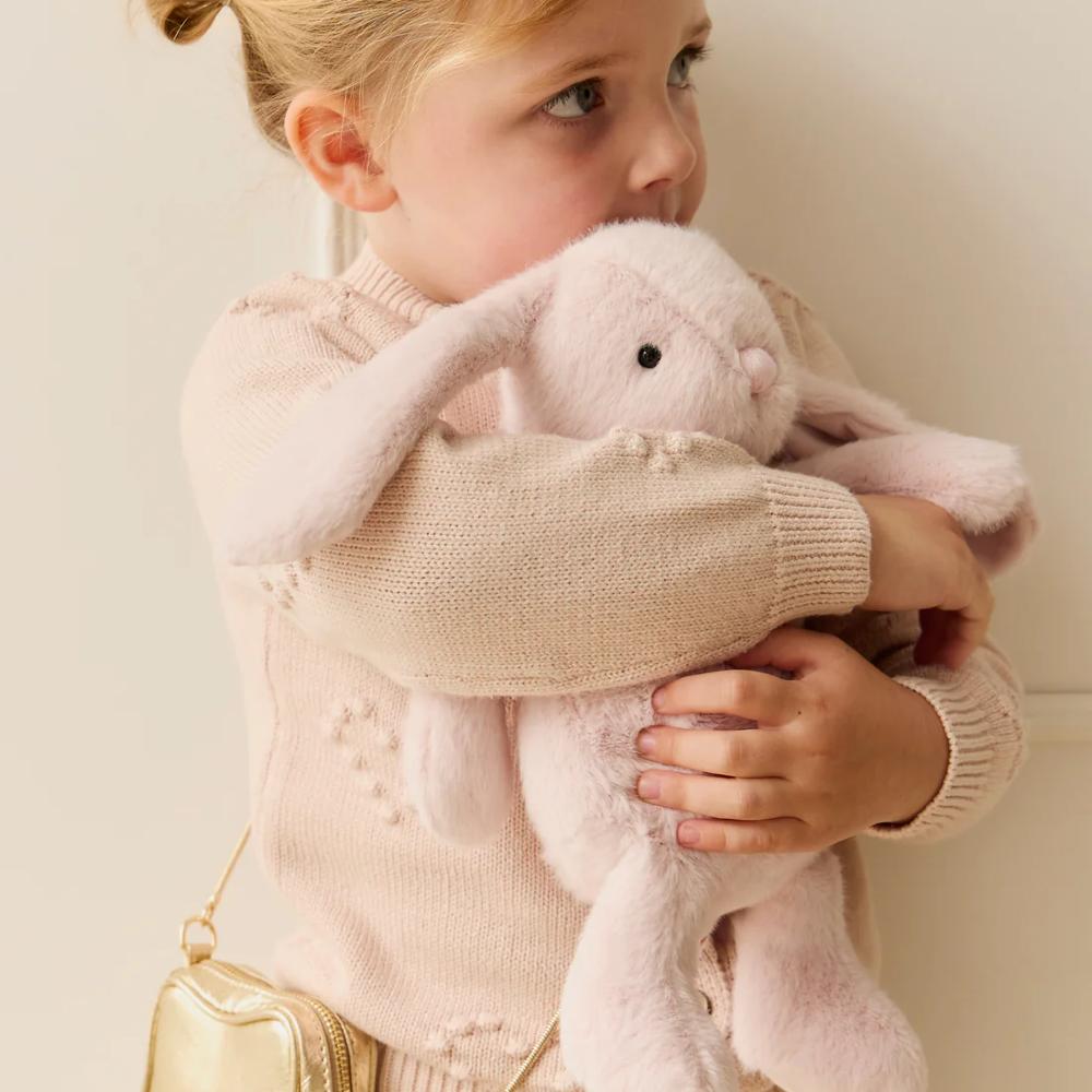 Baby holding light pink Jamie Kay bunny soft toy against white background.