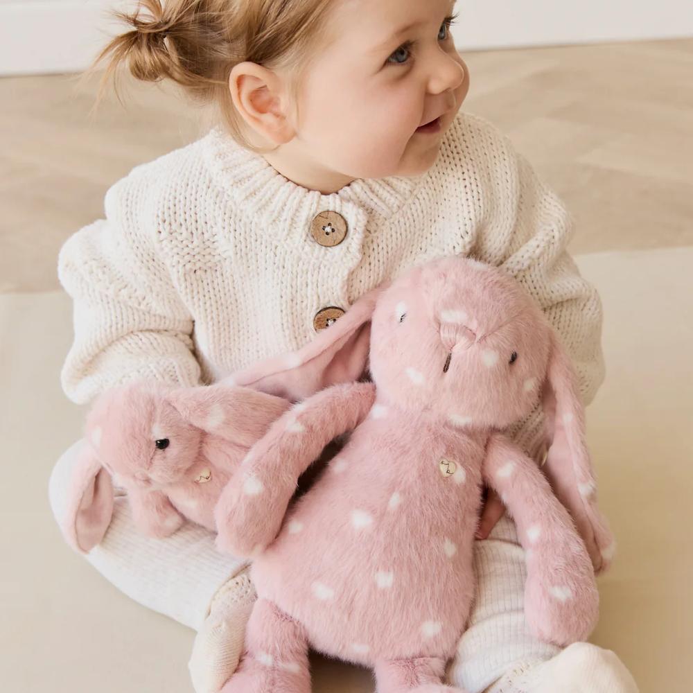 Baby holding Jamie Kay pink bunny soft toy sitting on floor.